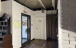 Лофт Повод Есть. Коридор с белыми кирпичными стенами, деревянным полом, стеклянной дверью с черной рамой, рядом вешалка с коричневыми занавесками. На стене желтая схема. Потолок с открытыми коммуникациями и висящими лампами.