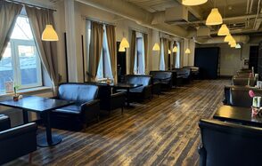 Лофт Повод Есть. Просторное помещение с деревянным полом, длинный ряд черных кожаных диванов и столиков, окна с занавесками, подвесные светильники с теплым светом, белые стены, вентиляционные трубы на потолке, цветы на столах.