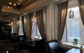 Лофт Повод Есть. Пространство с большими окнами, белыми стенами и подвесными светильниками. Расставлены кожаные диваны и кресла с темными столами. На подоконниках и столах цветы. Стеклянные двери ведут в соседнее помещение. Потолочные трубы и деревянный пол.