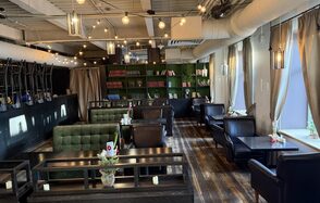 Лофт Повод Есть. Лофт-пространство с деревянным полом и подвесными светильниками. Зоны отдыха с зелеными диванами и черными креслами. Темные деревянные столы. Зеленый книжный стеллаж на заднем плане. Окна с бежевыми занавесками, подвесные лампы. Декорированы цветами.