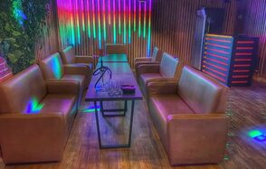 Forest(Лес). Пространство с деревянными стенами и полом, два ряда коричневых кожаных диванов напротив друг друга, стол посередине, кальян и стаканы на столе. На стене разноцветные светодиодные лампы. Угловая барная стойка с подсветкой.