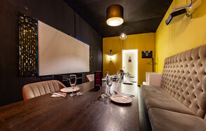 Rubin Loft (Рубин Лофт). Зал Гэтсби. Уютная комната с длинным деревянным столом и стульями, с одной стороны мягкий диван. Желтые стены, черный потолок. На стене большой экран и декоративные панели. Освещение: подвесные светильники и бра. Сервировка: бокалы, тарелки, приборы.