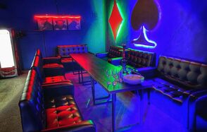 Rave(Рейв). Длинный стол в центре с черными кожаными креслами и диванами по периметру. Неоновое освещение: красный, зеленый, синий. На стене светящиеся символы карточных мастей. На столе набор бокалов и тарелок. Левый угол с холодильником.