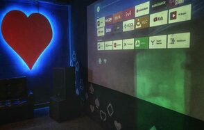 Rave(Рейв). Комната с проектором, на стене отображены иконки приложений. В левом углу большое красное сердце на синем фоне. Черная стена с белыми и красными фигурками в форме крестов и бубен. Справа темное кресло.
