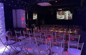 Лофт Репаблик. Темное помещение с рядами белых складных стульев, ориентированных на стены с проекторами. Черный потолок, яркие цветные световые эффекты. Справа виден диван. На стенах видны декоративные элементы белого кирпича.