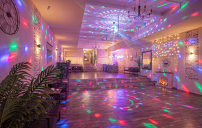 Loft Lodge (Лофт Лодж) 7. Просторный уютный лофт. Просторное помещение с кирпичными стенами, деревянным полом и разноцветным световым оформлением. Потолочные люстры, стулья и столы в задней части. Имитированный камин, настенные часы и растения по бокам. Текст на стене: "Let's PARTY!"