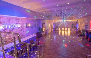 Loft Lodge (Лофт Лодж) 7. Просторный уютный лофт. Просторное помещение с кирпичными стенами и деревянным полом. Слева барная стойка и высокие стулья. Сверху подвесные светильники. В центре помещения диско-шар и мерцающие огни. На заднем плане большие световые буквы «LOVE».