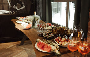 Площадка для мероприятий в стиле лофт. Деревянный стол с закусками и напитками: нарезанные фрукты, тарталетки, бокалы с напитками. Ваза с цветами. Окна с затемняющими шторами, стена с кирпичной отделкой. Паркетный пол, элементы в темных тонах, окна с дневным светом.