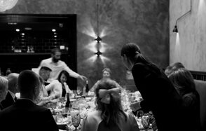 Площадка для мероприятий в стиле лофт. Частично затемнённое пространство с кирпичной стеной, чёрной плиткой на заднем плане, большим столом, за которым сидят люди. Настенные светильники, оформление в тёмных тонах, мебель для обеда или мероприятия.