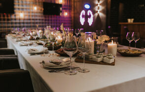 Площадка для мероприятий в стиле лофт. Пространство с длинным банкетным столом, покрытым белой скатертью. На столе сервировка из бокалов, тарелок, свечей и закусок. На стене неоновая вывеска. Тёплое освещение с настенными лампами. Темные стены и декоративные элементы.