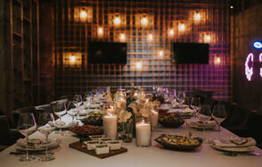 Площадка для мероприятий в стиле лофт. Длинный стол в зале с решетчатыми стенами, сервирован белой скатертью, украшен свечами и посудой. Потолочные светильники создают мягкое освещение. В центре стола блюда с едой. На стене два экрана и неоновая надпись.