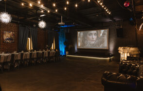 Nord Loft(Норд Лофт). Просторное праздничное помещение с кирпичными стенами и темным потолком. Длинный стол с белыми скатертями и стульями справа, кожаный диван слева. На стене большой экран с проектором. Подвесное освещение с гирляндами и декоративными светильниками.