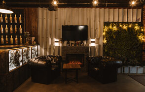 Nord Loft(Норд Лофт). Лофт-пространство с камином, два кожаных кресла, журнальный столик. На стене телевизор, рядом подсвеченные бутылки и живая стена с растениями. Освещение мягкое, с использованием подвесных ламп. Интерьер темный, с элементами из дерева и мрамора.