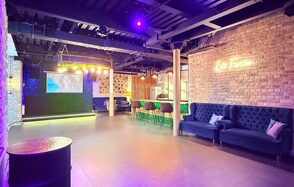 Лофт Фантом. Лофт-пространство с деревянными и кирпичными стенами; большая центральная зона с синими диванами; барная стойка с высокими стульями у стены; подсветка фиолетового оттенка; на стене надпись неоновыми буквами.