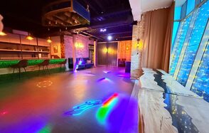 Лофт Фантом. Просторное помещение с танцполом и неоновым светом, барная стойка с высокими стульями, стол из светлого дерева, кирпичная стена, большие окна с гирляндами огней, черный потолок с балками, разноцветные световые узоры на полу.