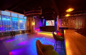 Лофт Фантом. Просторное помещение с деревянным баром, ряд разноцветных стульев, кирпичная стена с экраном, большое окно с вечерним видом и светодиодной подсветкой, разноцветное освещение, темный потолок, светящиеся столы у окна, колонна и декоративная перегородка.