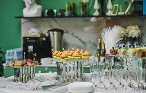 Arcobaleno Loft(Аркобалено Лофт). Праздничное пространство со столом для фуршета. На столе бутерброды, закуски в стеклянных стаканах, десерты, стеклянные бокалы. На заднем плане декор и напитки, зеленые стены, бело-мраморная столешница.
