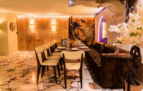 Стильный лофт Джаванди. Современное помещение с мраморным полом. Длинный стол с сервировкой и восемью стульями с бежевыми сиденьями. Справа диван, в углу ваза с белыми цветами. На стене деревянные панели и декоративная картина. Мягкое освещение.