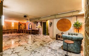 Стильный лофт Джаванди. Просторное помещение с мраморным полом, барной стойкой и высокими стульями. Голубой диван и круглый столик. Настенные декоративные элементы с оранжевыми кругами. Украшено растениями. Потолочные светильники и проектор. Стены светло-коричневые.