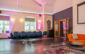 Просторный лофт с верандой и внутренним двориком. Просторная комната с высоким потолком и белыми кирпичными стенами, освещенная разноцветными светодиодами. Длинный стол с темно-синими стульями, оранжевый диван и ковёр. Окна с жалюзи, два круглых столика, зеркало и декор на стенах.