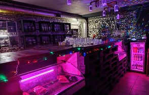 Кинолофт. Подвальное помещение с барной стойкой, оформленной темными паллетами и стеклянной поверхностью, подсветка разноцветными гирляндами. На полу плитка. Задняя стена декорирована черно-белыми изображениями. На потолке подвесные светильники. Справа холодильник с напитками.