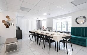 Современный зал с зеркалами. Просторное светлое помещение с длинным столом, накрытым светлой скатертью, и черными стульями. На стене круглое зеркало и шестигранные декоративные элементы. Напольное покрытие — светлая плитка. Прямоугольные окна с видом на улицу.