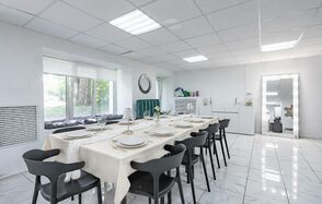 Handy Class (Хэнди Класс). Современный зал. Современное светлое помещение с длинным столом, накрытым белой скатертью, и черными стульями. Окно с видом на природу, белые стены и плиточный пол. Зеркало на стене, настольные лампы, декор в виде растений, потолочные светильники.
