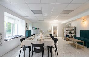 Handy Class (Хэнди Класс). Современный зал. Просторная комната с белым плиточным полом и подвесным потолком. Длинный обеденный стол с черными стульями, накрыт скатертью. Диваны изумрудного цвета, стеллаж с декором и зеркальная лампа. Окно с подоконником и сиденьями.