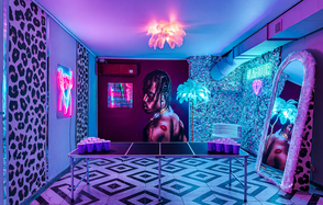 Неоновый лофт Гламур. Квадратная комната с шахматным полом, неоновыми вывесками, зеркалом в яркой раме и большим постером на стене. В центре стол с пластиковыми стаканами. Освещение розово-фиолетовое, на потолке светильник, похожий на перья.
