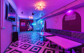 Неоновый лофт Гламур. Развлекательное пространство с геометрическим полом, фиолетовым освещением и неоновыми вывесками. Длинный серый диван вдоль стены, два прямоугольных стола. Декоративные светильники на потолке, настенные изображения и растения с подсветкой.