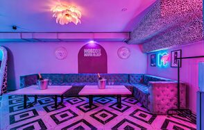 Неоновый лофт Гламур. Караоке-зал с угловыми диванами обития, два стола с ведёрками шампанского, плиточный пол с геометрическим рисунком, неоновая надпись, яркая люстра, фиолетово-розовая подсветка, леопардовый принт на потолке, круглые зеркала, декорированные стены.