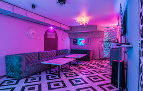 Неоновый лофт Гламур. Караоке-зал с неоновой подсветкой, фиолетовые и розовые световые акценты. Угловой диван из зелёного бархата, два прямоугольных стола, черно-белый узорчатый пол, стены с декоративными элементами и телевизор. Табличка с надписью и неоновый логотип.