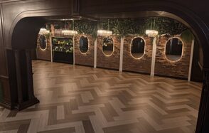 Зал с кухней до 150 гостей. Пространство с паркетным полом ёлочкой, кирпичными акцентными стенами с овальными зеркалами и подвесными светильниками. Плотные зелёные растения вдоль верхней части стен. Тёмная деревянная отделка потолка и арки.