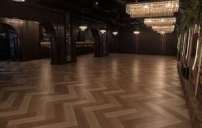 Зал с кухней до 150 гостей. Просторный зал с паркетным полом, уложенным елочкой. На потолке висят большие хрустальные люстры, создающие мягкое освещение. Стены темные, декорированы зелеными растениями и кирпичом. Вдоль стены барная стойка.