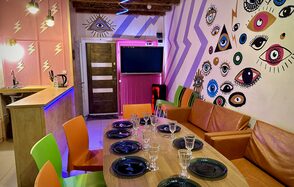 Яркий двухуровневый лофт. Накрытый посудой стол. Разноцветные стулья. Диваны. Барная стойка. Разукрашенные рисунками стены разноцветные, яркие стены. Телевизор