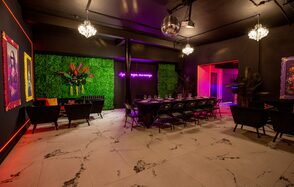 Белый неоновый лофт-зал. Торжественное пространство с зелёной растительной стеной, длинным столом и стульями. Потолок тёмный, на нём зеркальный шар и люстры. Светлые мраморные полы. На стене неоновая надпись и картины. Освещение разноцветное.