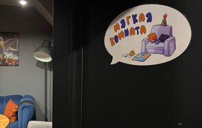 Мягкая комната на 6 гостей. Часть комнаты с серыми стенами, на стене постер. Диван синего цвета с оранжевыми подушками. Большой торшер с белым абажуром. На двери наклейка с иллюстрацией кресла и текстом «Мягкая комната».