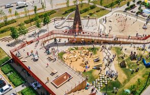 Площадка c одной фотографией в Москве. Детская площадка с деревянным настилом, песочницей, горками и лазалками. Центральная конусообразная конструкция с несколькими спусками. Зеленые насаждения и дорожки вокруг. Люди гуляют по территории. Площадка огорожена забором.