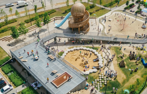 Площадка c одной фотографией в Москве. Открытое детское площадное пространство с деревянной башней и винтовой горкой, песочницей, зонами отдыха и дорожками. Терраса с кафельным покрытием и столиками. Разноцветные игровые элементы разбросаны по зеленому газону.