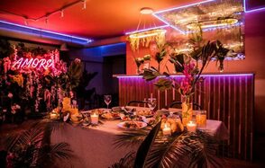 Тропический лофт с современным дизайном и верандой. Праздничное пространство с сервированным столом на шесть персон, украшено зеленью и свечами. Неоновая надпись "AMOR", яркое освещение синими и красными тонами, подвесные светильники. Стены с деревянными панелями, потолок украшен зеркальной плиткой.