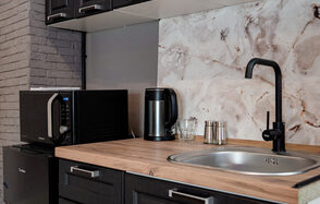 Proloft(Пролофт) . Кухонный уголок с раковиной из нержавеющей стали и черным краном, деревянная столешница, на ней черная микроволновка, металлический чайник и стеклянные стаканы. Задняя стена отделана серыми кирпичами и мраморной плиткой. Черные шкафы с серебристыми ручками.