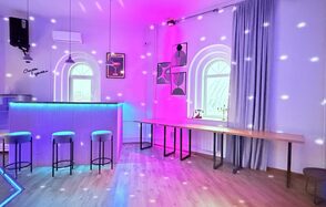 Центрум. Уютное светлое пространство круглой формы. Барная зона с подсветкой, высоким столом и тремя стульями. Деревянный пол, белые стены с арочными окнами. Подвесные светильники, дискотечное освещение. Длинный стол у стены, постеры, серые занавески, кондиционер на стене.