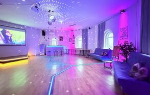 Центрум. Уютное светлое пространство круглой формы. Просторное помещение с диджейским пультом и экраном, деревянный пол, неоновая подсветка, дискошар на потолке, два синих дивана, арочные окна, высокий стол с табуретами, цветные светильники, украшения на стенах, растения.