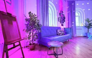 Центрум. Уютное светлое пространство круглой формы. Современное пространство с деревянным полом и сиреневыми стенами. Синий диван с подушкой, рядом кофейный столик, мольберт и искусственные растения. Арочные окна, розовое освещение, настенные фотографии в черных рамках. Шторы закрывают некоторые окна.