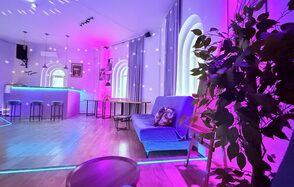 Центрум. Уютное светлое пространство круглой формы. Пространство с фиолетовой подсветкой, барная стойка с высокими стульями, деревянный пол, диван с подушками, столики, арочные окна с занавесками, декоративное растение, подвесные светильники, потолочный свет.