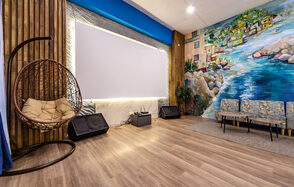 Rubin Loft (Рубин Лофт). Зал Сицилия. Помещение с деревянным полом, висячим плетеным креслом, светлой стеной с крупным экраном, двумя динамиками, мягким диваном. Одна стена украшена ярким настенным рисунком с изображением побережья.