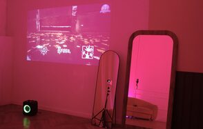 Стильный лофт для уютных вечеринок. Проекционный экран на стене с изображениями и текстом. Напольное зеркало с деревянной рамой. Колонка на полу. Розовое освещение, паркетный пол, светлый диван за углом. Камера на штативе.