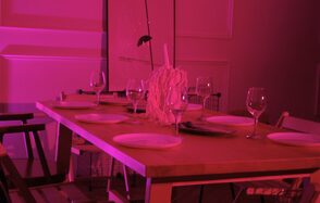 Стильный лофт для уютных вечеринок. Прямоугольный деревянный стол с расставленными белыми тарелками и бокалами для вина. В центре стола декоративная композиция. Окружают разностильные стулья. Освещение розового цвета, стены с белыми панелями.