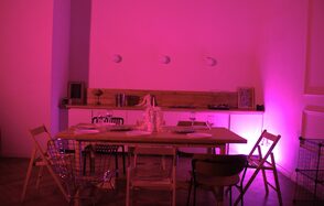 Стильный лофт для уютных вечеринок. Столовая зона с деревянным столом, окружённым разнообразными стульями. На столе белая салфетка и бокалы. Позади небольшая барная стойка с предметами декора и освещением розового тона. Пол паркетный, на стене три светильника.