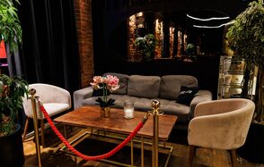 Двухуровневая event-площадка 550 м². Лаунж-зона с серым диваном и двумя белыми креслами вокруг деревянного стола, украшенного цветочным горшком и свечой. Красная веревка на золотых стойках. Зелёные растения по краям. Темные занавески и кирпичная стена. Зеркало на стене.